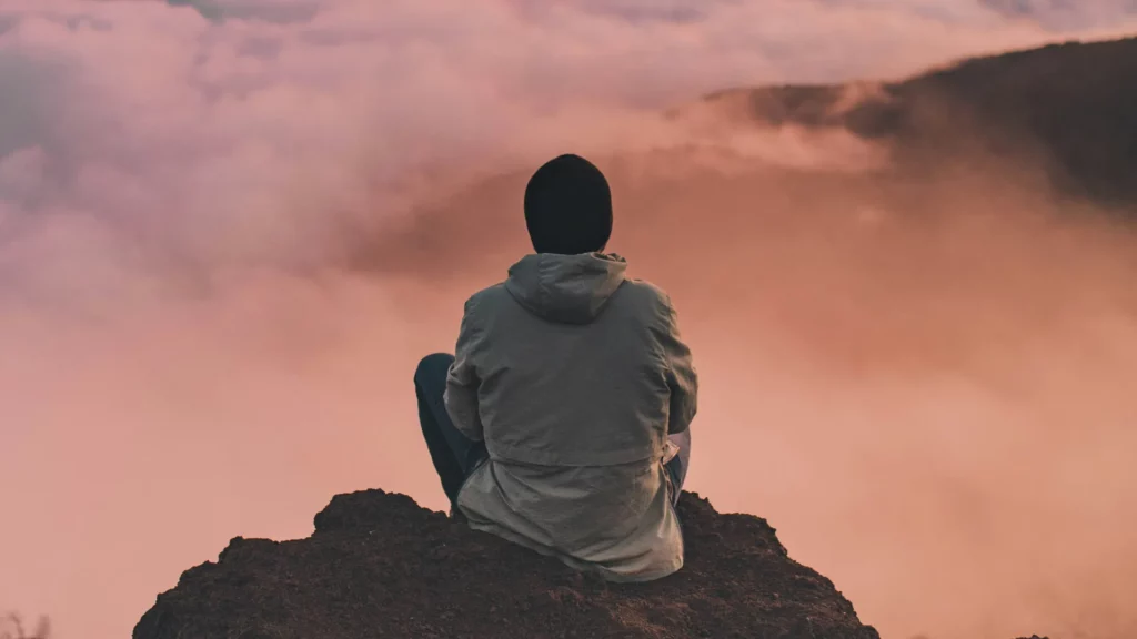 Personne assise au sommet d'une montagne au-dessus d'une mer de nuages, au coucher du soleil — reprendre son souffle face au burnout, hypnose à Lausanne